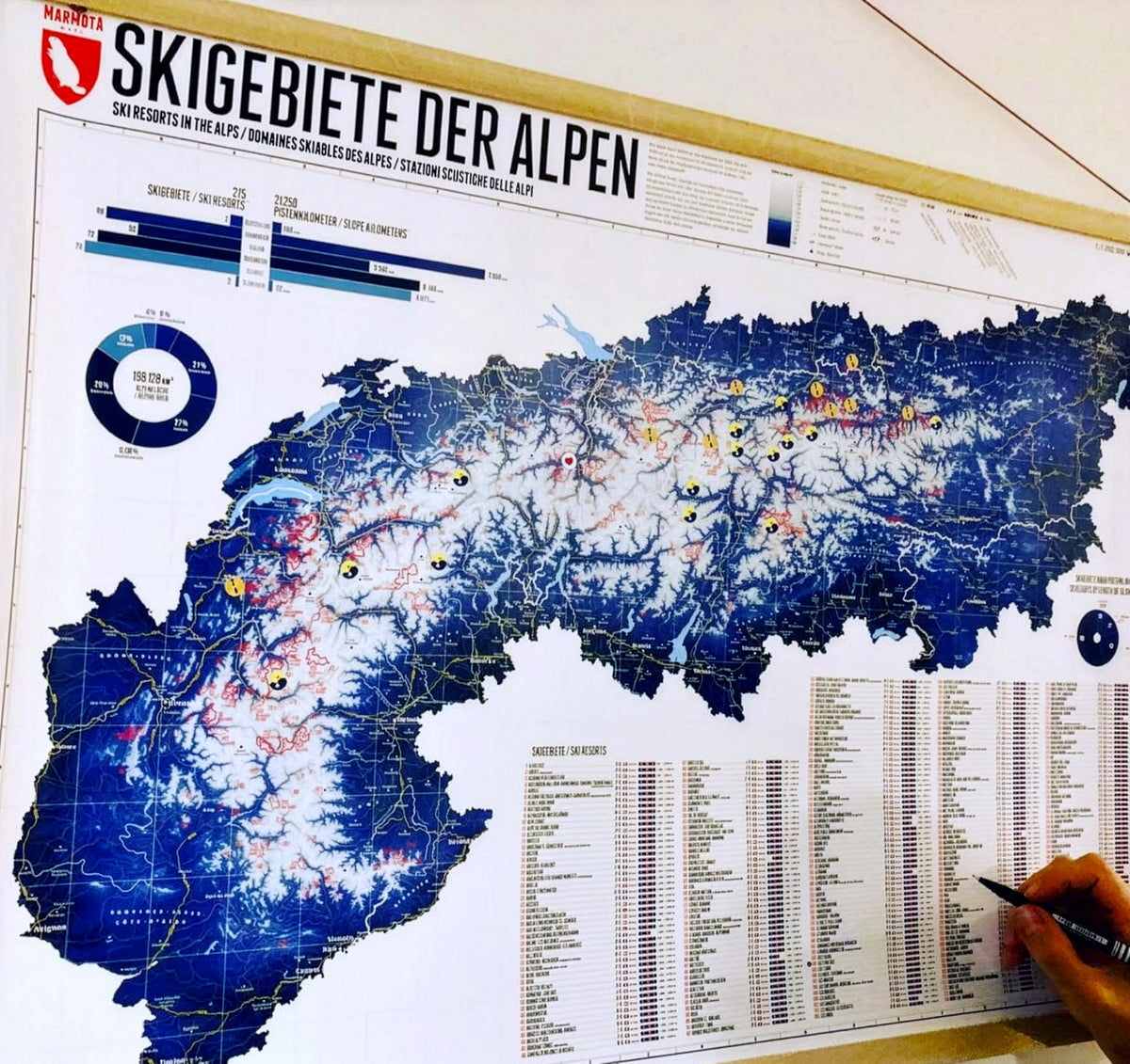 Alpenkarte mit 263 Skigebieten – an der Wand, eine Hand markiert Skigebiete in der Liste, große Ansicht